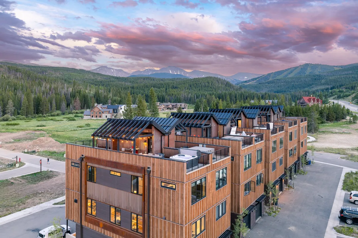 The image showcases a striking, modern single-family house set against a spectacular mountain backdrop at sunset. The architecture is sleek and inviting, with expansive glass windows and multiple outdoor seating areas. The sky glows with warm sunset colors, while cozy fire pits and lounge spaces hint at relaxing evenings spent outdoors. The scene evokes both tranquility and excitement, offering sweeping views and a sense of upscale mountain living.