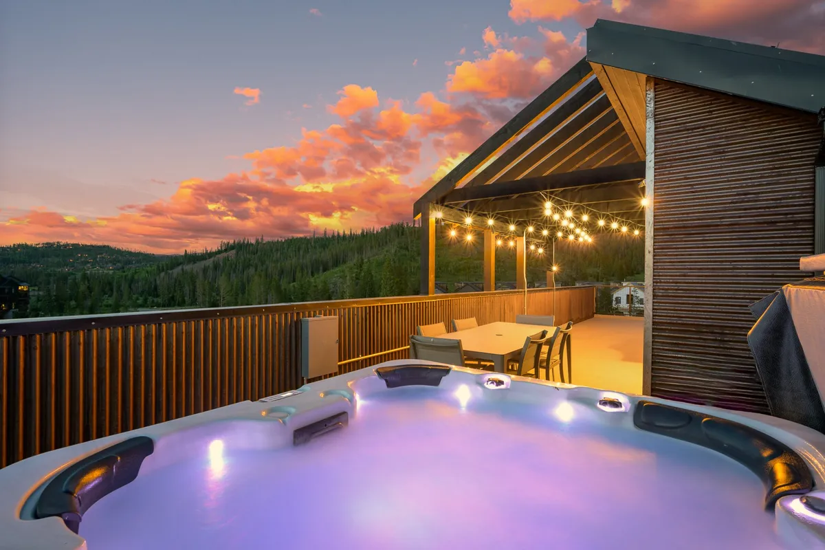 A tranquil outdoor patio scene with a bubbling hot tub, spacious dining table, and twinkling string lights. The area overlooks a breathtaking mountain range, ideal for entertaining or unwinding after a day of adventure.