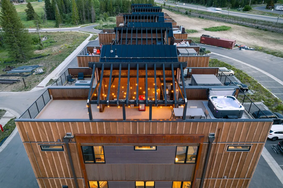 A vibrant rooftop terrace featuring a hot tub, comfortable outdoor seating area, and glowing string lights overhead. The terrace is set against a backdrop of expansive mountain views, creating an inviting space for socializing, relaxing, or enjoying a sunset meal.