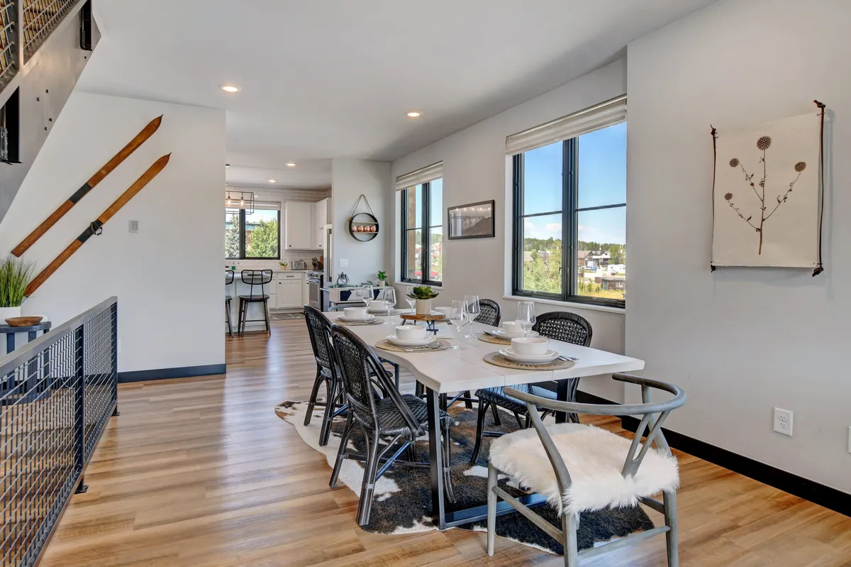 A dining area with a substantial live-edge wood table set for a meal, large windows allowing in warm sunlight, and decorative elements like skis on the wall, evoking a sense of place and celebration. The space is arranged for family-style or group dining.