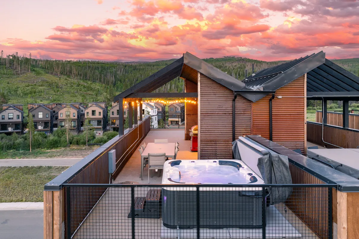 A cozy exterior shot featuring a hot tub, a welcoming outdoor seating area, string lights overhead, and scenic mountain views. The setup invites relaxation and socialization, perfect for outdoor dining or stargazing in the evening with family and friends.