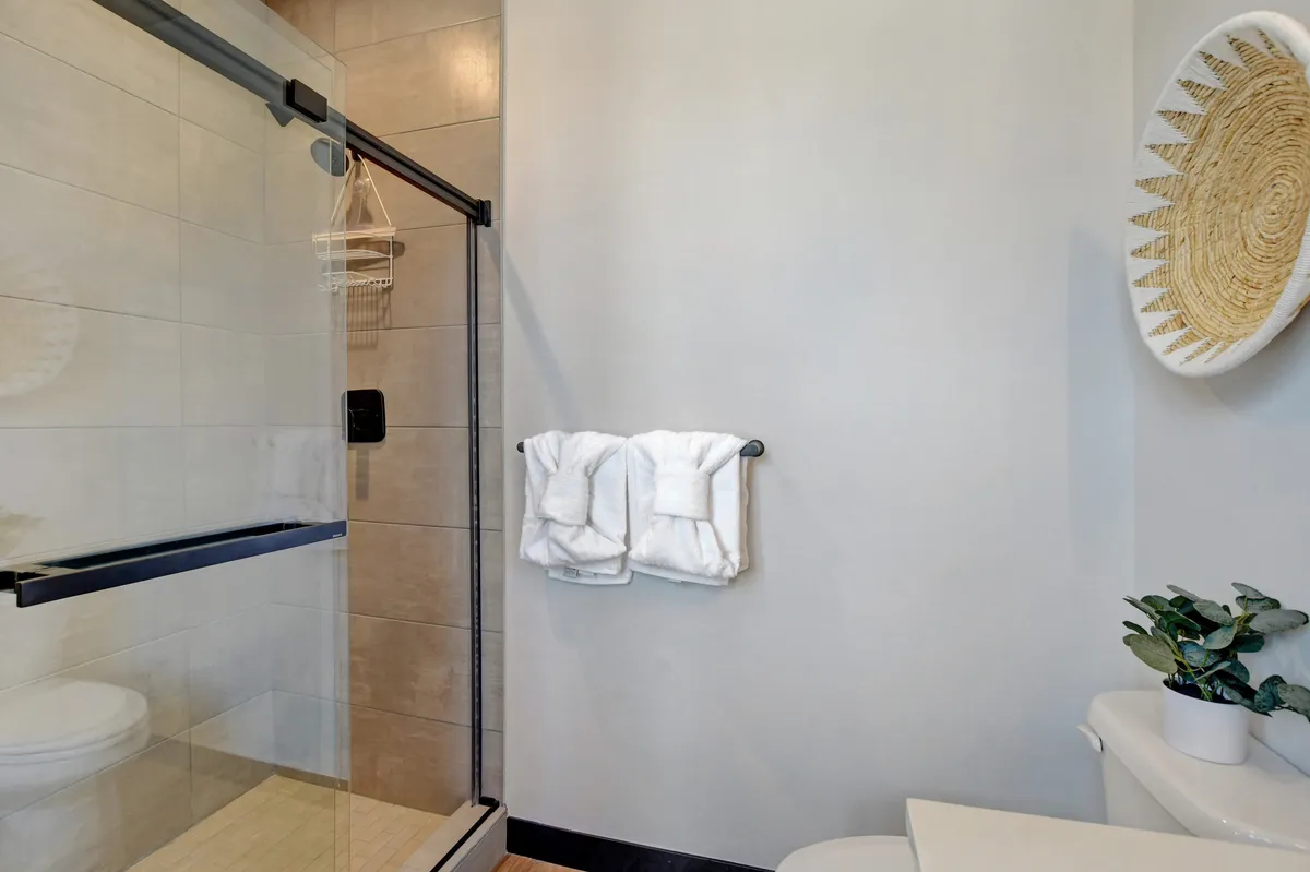 A bright, spa-inspired bathroom with a glass shower enclosure, neatly stacked towels, a decorative wall basket, and a touch of greenery for a tranquil feel. The space is clean, modern, and inviting—ideal for self-care and relaxation.