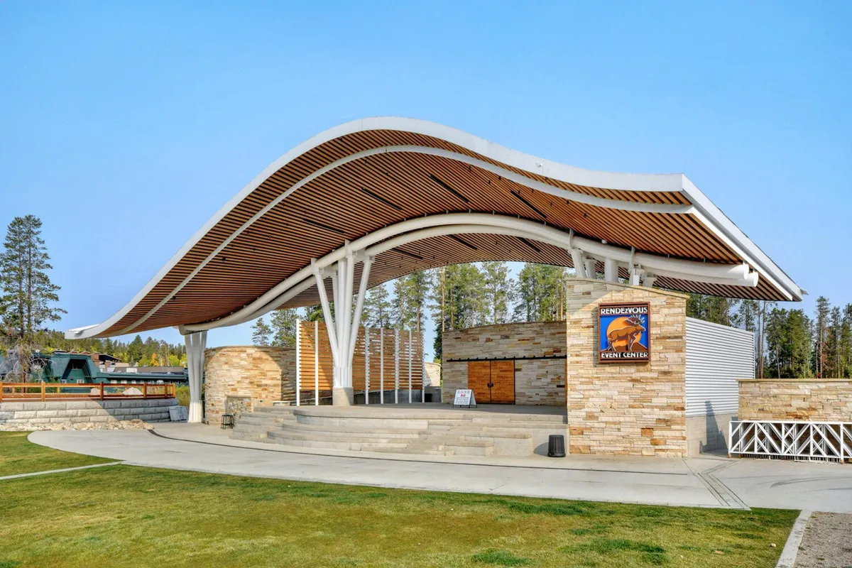An exterior shot of Hideaway Park's signature curved roof stage, stone facade, and open gathering area, evoking the energy of downtown. The scene hints at outdoor concerts, festivals, and community events under the mountain sky.