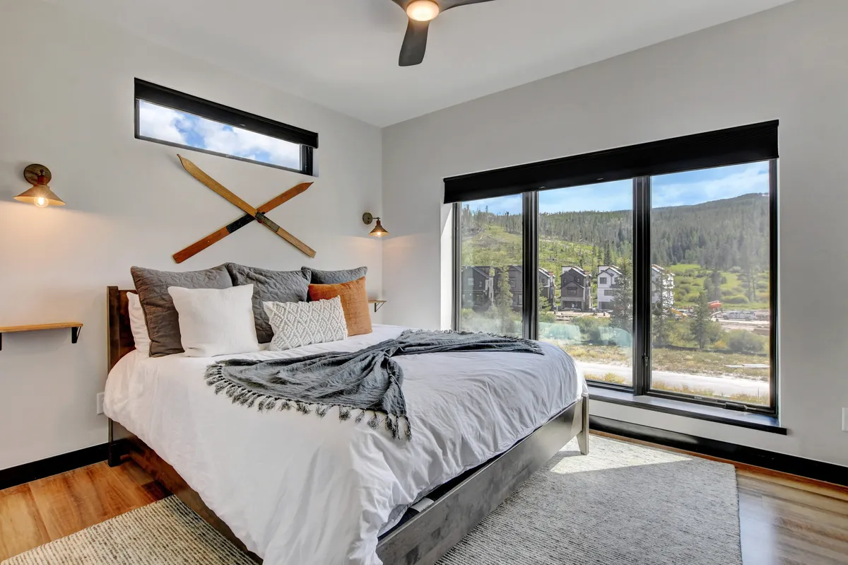 A spacious bedroom with a large bed positioned near expansive windows, offering mountain views and ample natural light. Decorative wall elements add character, while the cozy setup invites relaxation and reading.