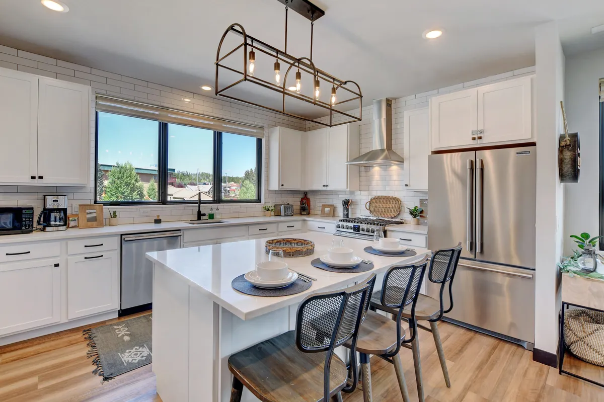 A bright, spacious kitchen featuring a large central island with bar seating, stainless steel state-of-the-art appliances, custom lighting, and expansive windows that flood the space with natural light. The kitchen island is a focal point, ideal for casual dining or gathering while prepping meals.
