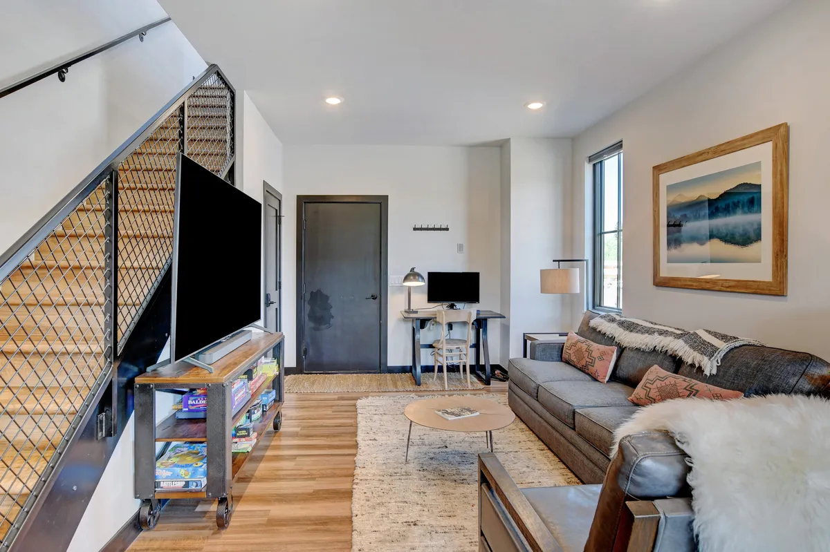 A versatile living space featuring a cozy sofa, a TV for movie nights, a staircase leading to another level, stylish artwork, and a compact desk for creative or work activities. The design encourages both relaxation and social interaction.