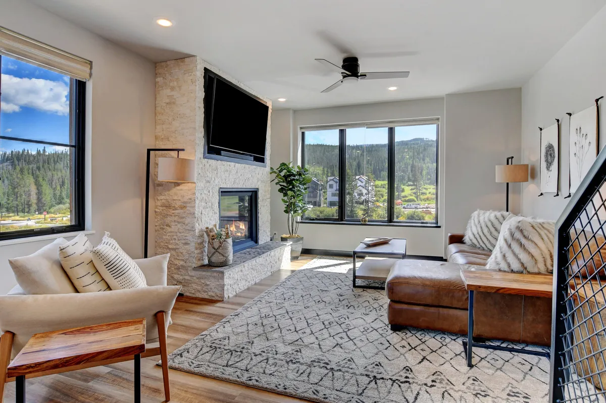 This image showcases a spacious, modern living room with a plush sofa centered around a custom gas fireplace. Large windows bathe the room in natural light and offer scenic views, while an area rug and potted plant add warmth and character. The setup is perfect for relaxing, reading, socializing, or watching TV.