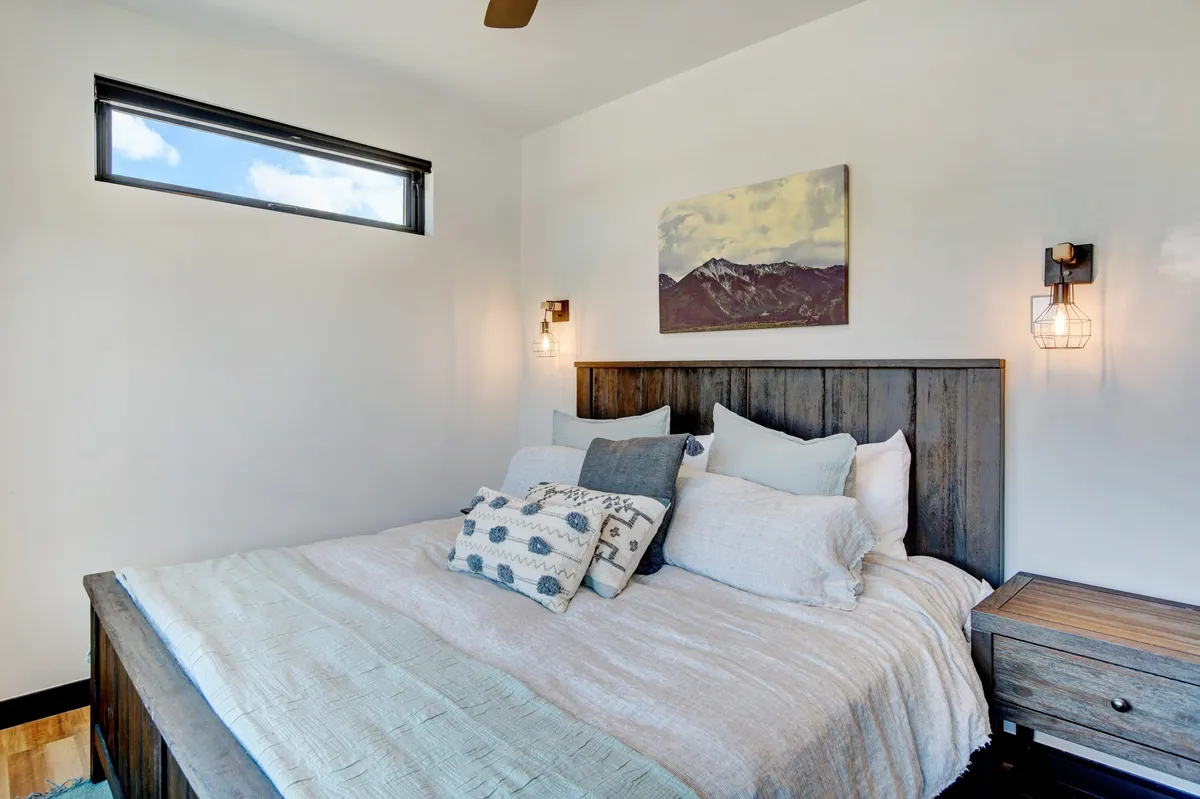 A tranquil bedroom featuring a plush bed with premium bedding, decorative pillows, and tasteful wall art. Natural light streams in from a nearby window, casting a warm, inviting glow over the serene space. A bedside table completes the restful ambiance, emphasizing comfort and relaxation.