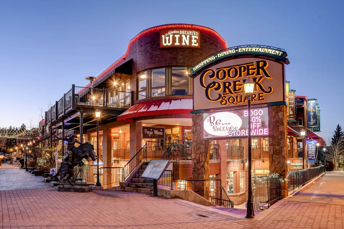 The image displays Cooper Creek Square in downtown Winter Park, featuring lively signage for gourmet dining, outdoor seating areas, and a community-focused sculpture. The setting is festive and bustling, alluding to local entertainment, shopping, and dining options within walking distance.
